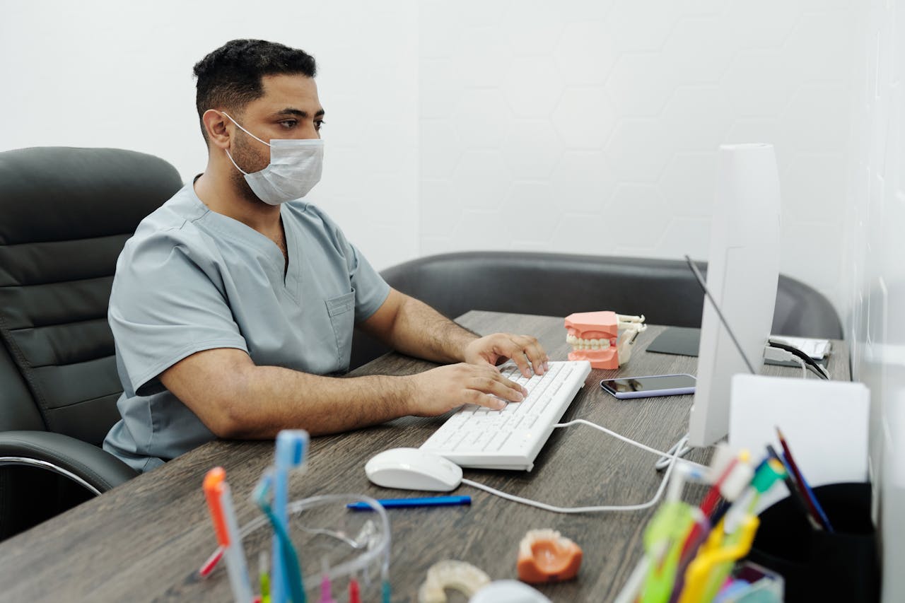 Dentist wearing mask working on computer at office desk with dental model.